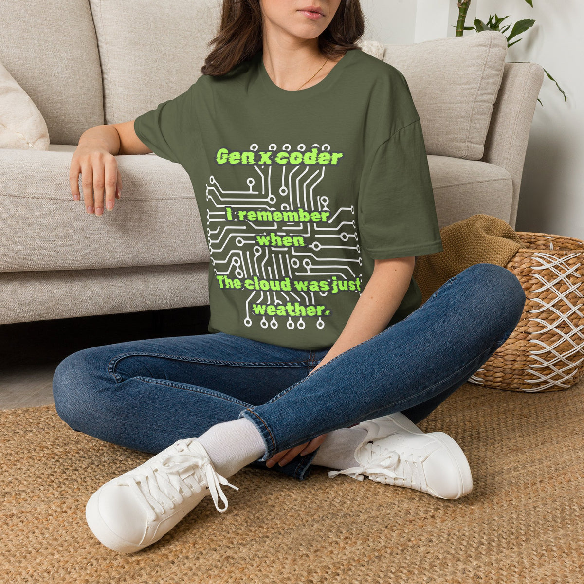 A woman sitting cross-legged on a woven rug in front of a beige sofa, wearing a green graphic t-shirt with the text 'Gen x coder I remember when The cloud was just weather paired with blue jeans and white sneakers.