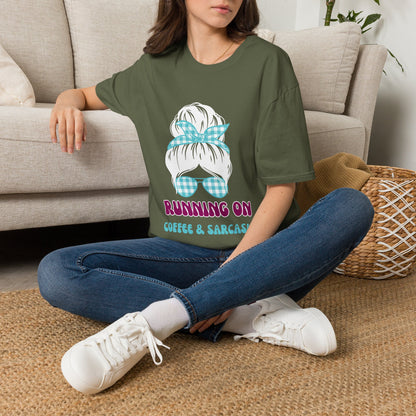A green short-sleeve graphic t-shirt displayed on a gray wood-plank background, featuring a cartoon illustration of a woman with a plaid hair bow and sunglasses, with the text 'RUNNING ON COFFEE & SARCASM'. 