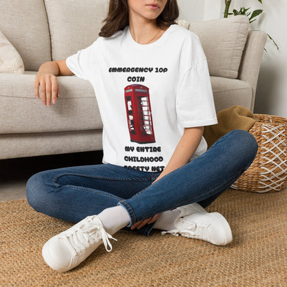 A woman sitting cross-legged on a woven rug in front of a beige sofa, wearing an oversized white T-shirt with the word 'EMMERGENCY 10P COIN, MY ENTIRE CHILDHOOD SAFETY NET' with a red telephone box graphic, blue jeans, and white sneakers.