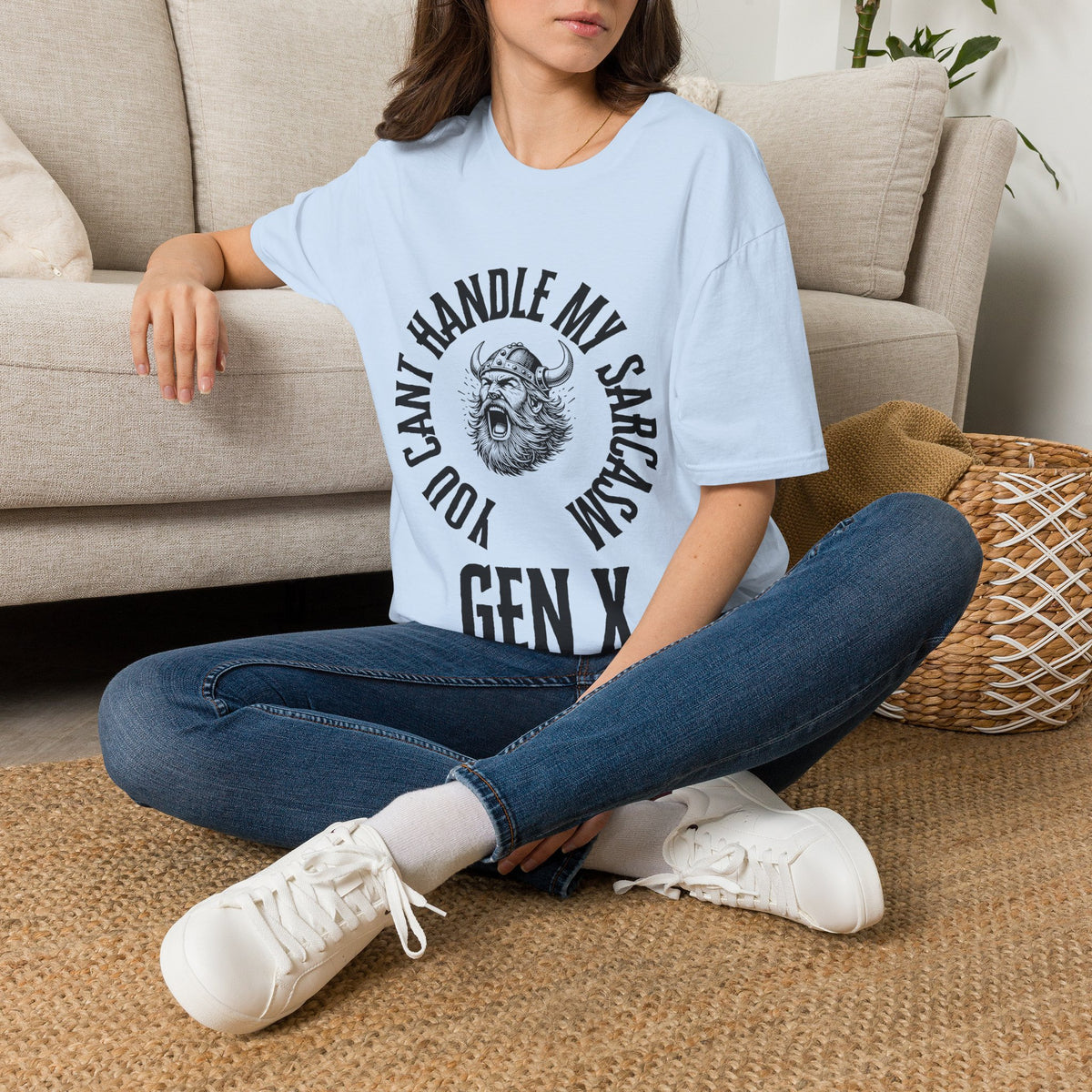 A woman sat on a tug next to a couch wearing a light blue short-sleeve graphic t-shirt with a black circular print featuring a roaring viking man and the text 'YOU CAN'T HANDLE MY SARCASM GEN X'. 