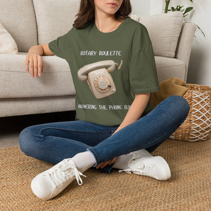 A woman sitting on a woven rug in front of a beige sofa, wearing a green graphic t-shirt with a vintage rotary phone design and the text 'ROTARY ROULETTE' and 'ANSWERING THE PHONE BLIND.
