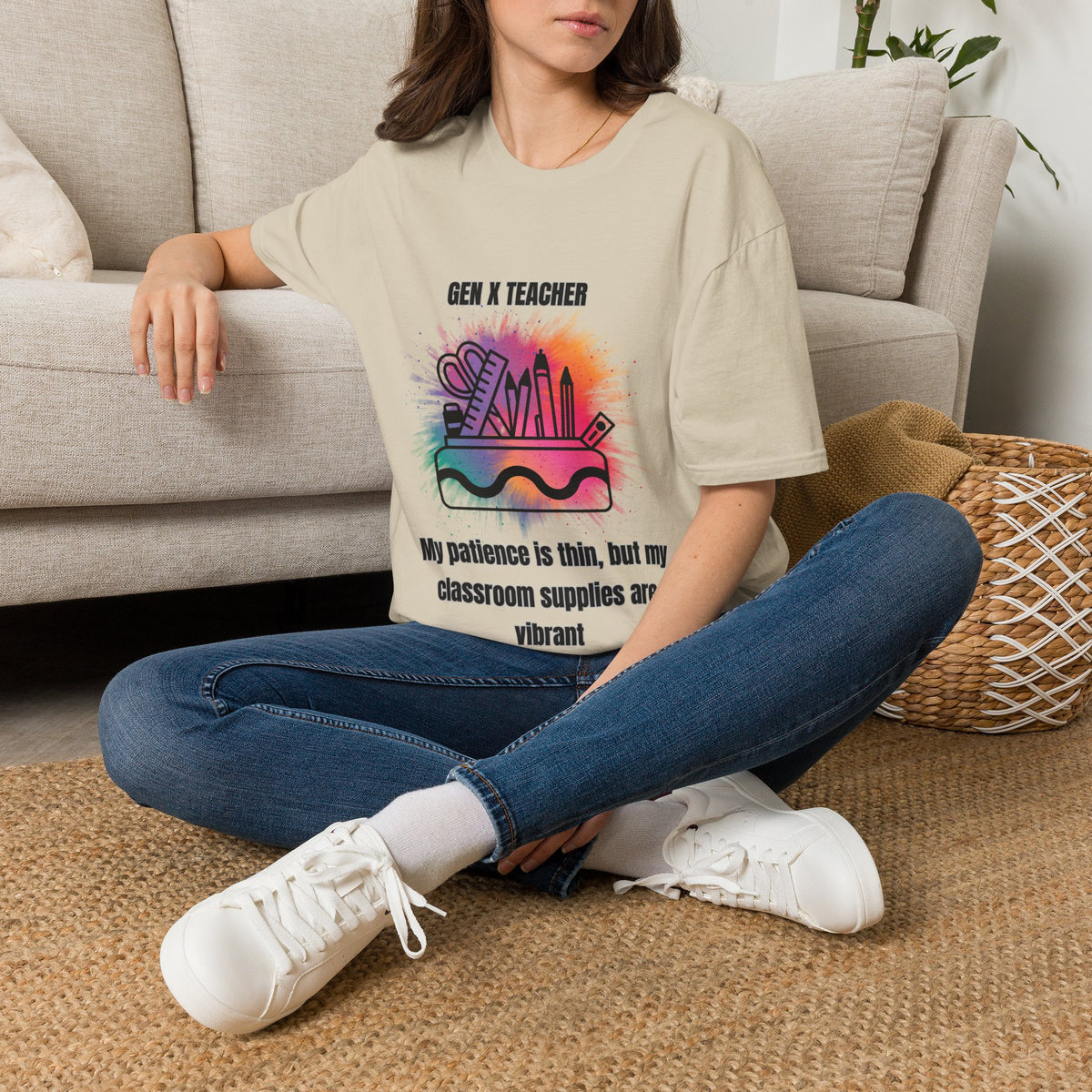 A woman sits cross-legged on a woven rug in front of a beige sofa, wearing an oversized beige graphic T-shirt with the text 'GEN X TEACHER' and a colorful school-supplies illustration, paired with blue jeans and white sneakers.