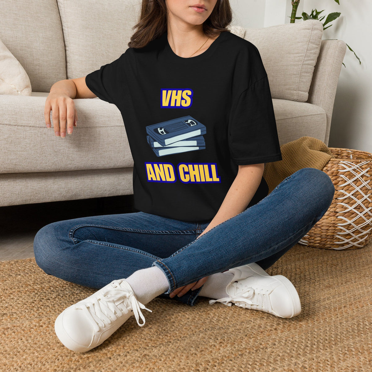 A woman sat on a rug next to a couch wearing a black graphic t-shirt, featuring a 'VHS AND CHILL' design with two VHS tapes. 