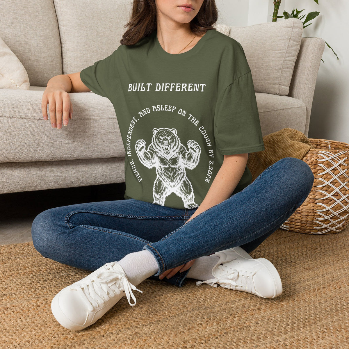 A woman sitting on a woven rug in front of a beige sofa, wearing a green graphic t-shirt with a white bear illustration and the text 'BUILT DIFFERENT'.