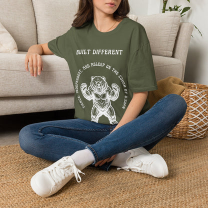 A woman sitting on a woven rug in front of a beige sofa, wearing a green graphic t-shirt with a white bear illustration and the text 'BUILT DIFFERENT'.