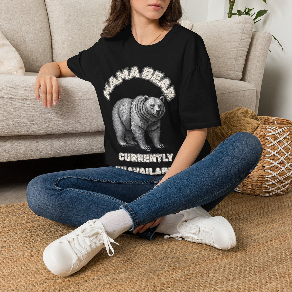 A woman sitting cross-legged on a woven rug in front of a beige sofa, wearing a black graphic T-shirt with the text 'MAMA BEAR' above and 'CURRENTLY UNAVAILABLE' below. paired with blue jeans and white sneakers.