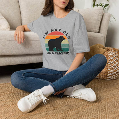 A woman sitting cross-legged on a woven rug in front of a beige sofa, wearing a gray graphic T-shirt with a bear silhouette and the text 'I'M NOT OLD I'M A CLASSIC' over a retro sunset design, paired with blue jeans and white sneakers.