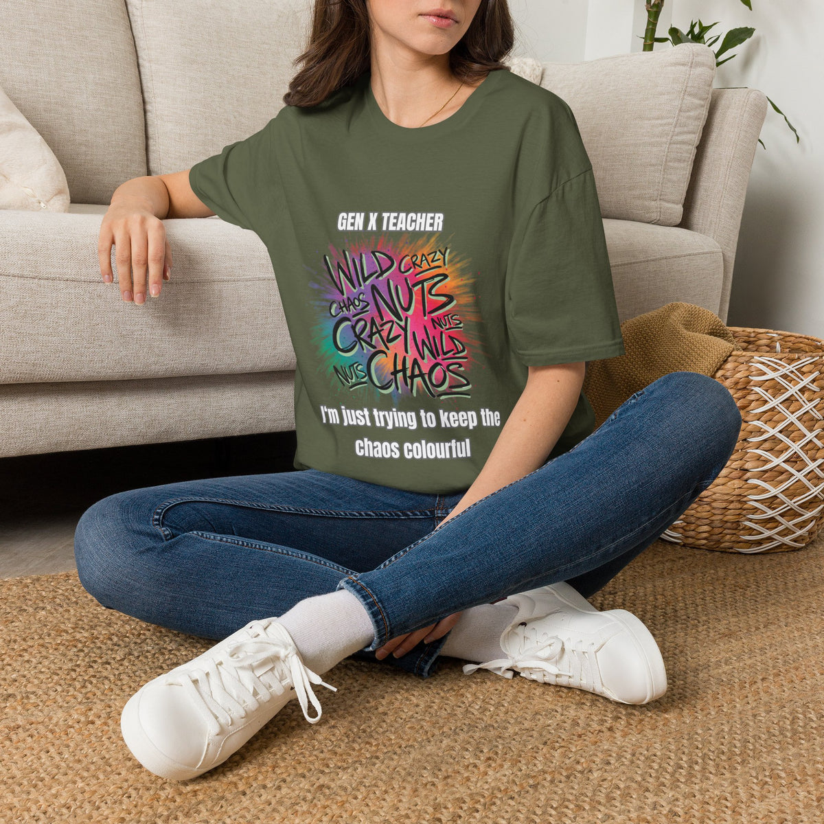 A woman sits cross-legged on a woven rug in a living room setting, wearing a green graphic t-shirt with colorful text that reads 'GEN X TEACHER' and 'I'm just trying to keep the chaos colourour!'. She is also wearing blue jeans and white sneakers.