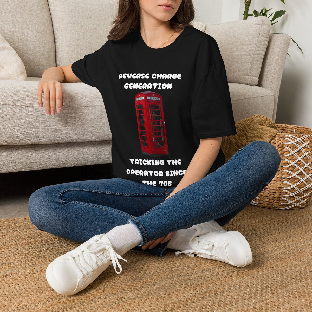 A woman sitting cross-legged on a woven rug in a living room setting, wearing a black graphic t-shirt with white text and a red telephone booth graphic, paired with blue jeans and white sneakers.