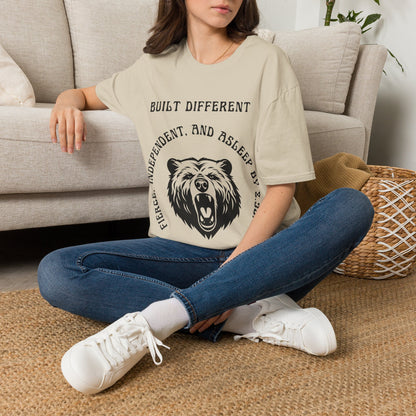 A woman sits cross-legged on a woven rug in front of a light beige sofa, wearing a sand graphic T-shirt with a black bear design and the text 'BUILT DIFFERENTShe is also wearing blue jeans and white sneakers.
