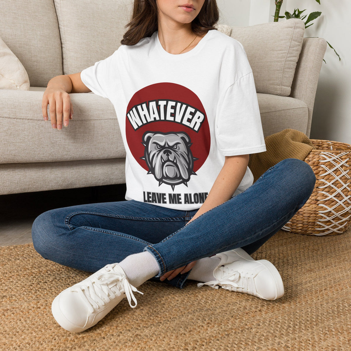A woman sitting cross-legged on a woven rug in a living room, wearing a white graphic t-shirt with a red circle, bulldog illustration, and the text 'WHATEVER LEAVE ME ALONE'.