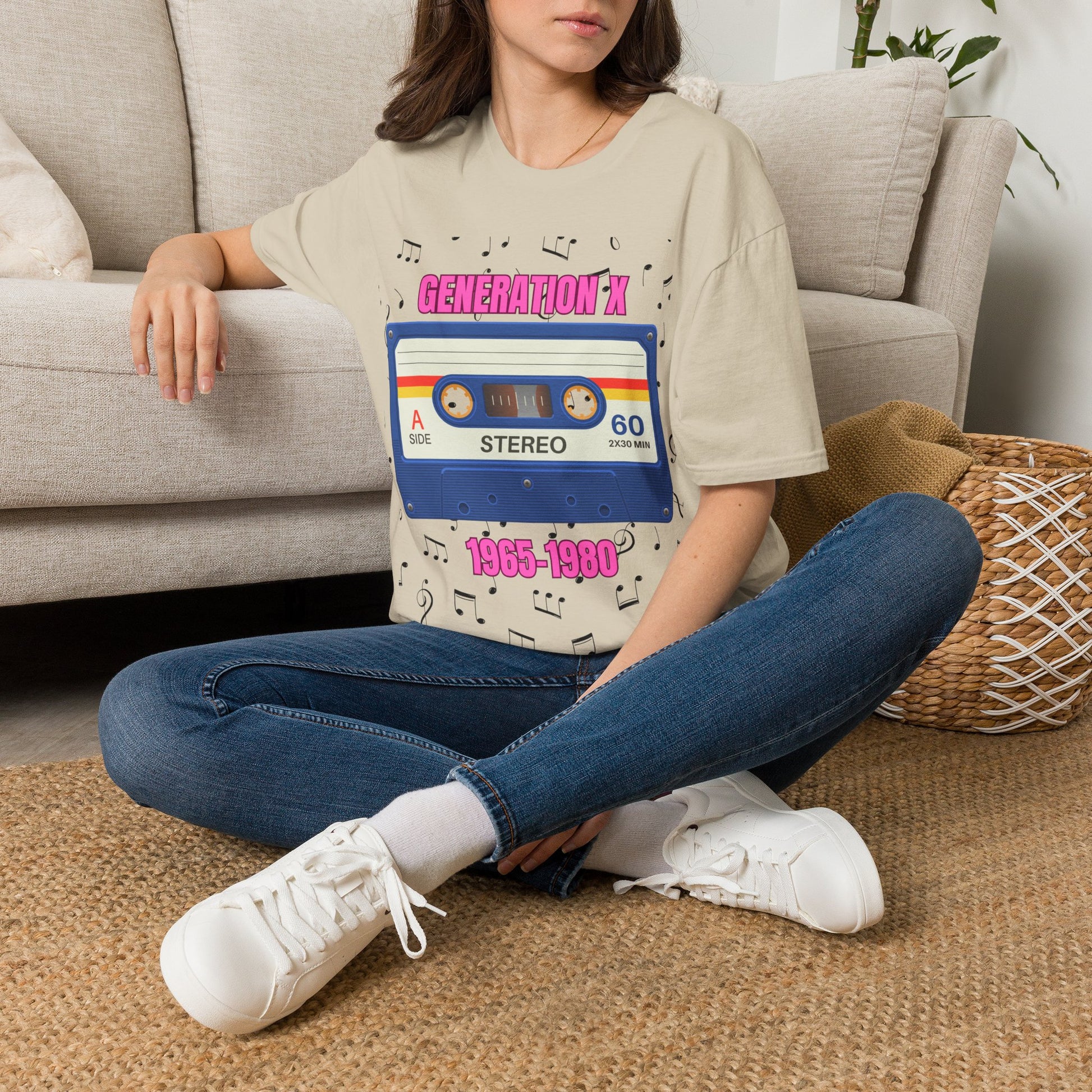 A woman sitting cross-legged on a woven rug in front of a light beige sofa, wearing a sand graphic T-shirt with a retro cassette tape design and pink text "GENERATION X 1865-1980" surrounded by music notes and blue jeans.