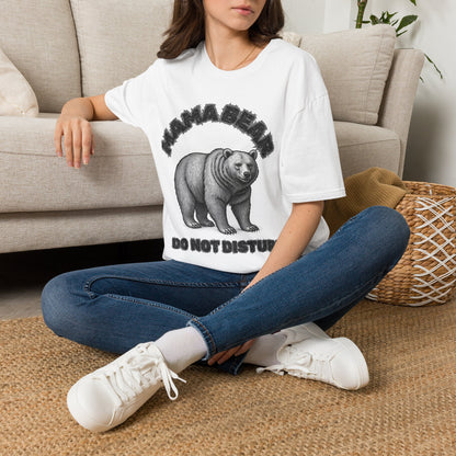 A woman sitting cross-legged on a woven rug in front of a beige sofa, wearing a white graphic T-shirt with a black bear illustration and the text 'MAMA BEAR DO NOT DISTURB'.