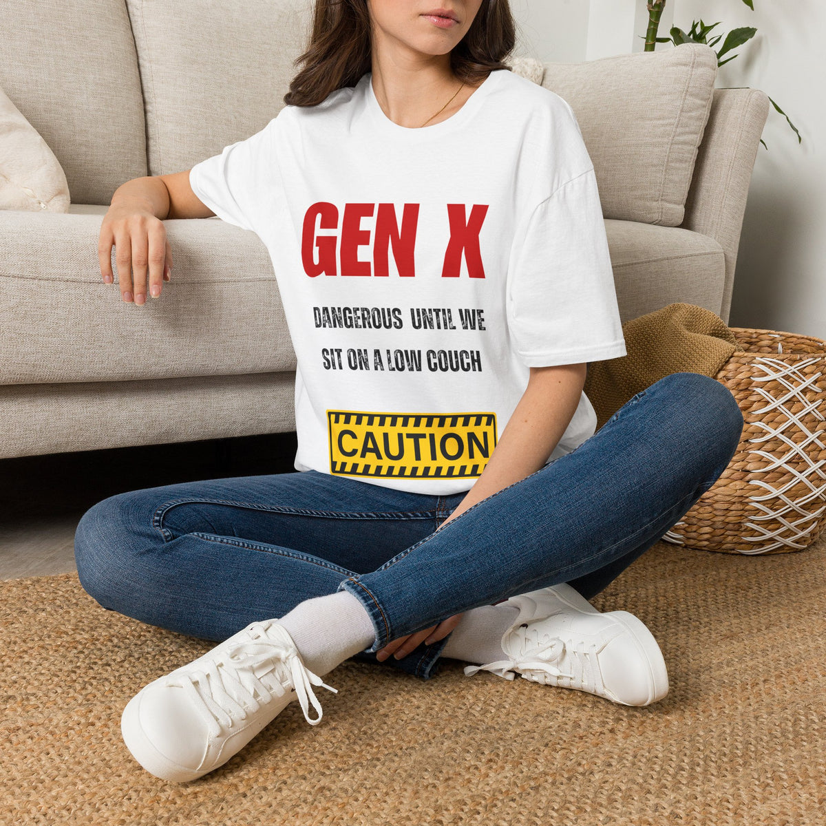 A woman sitting on a woven rug in front of a beige couch, wearing a white graphic T-shirt with red and black text reading 'GEN X DANGEROUS UNTIL WE SIT ON A LOW COUCH' and a yellow caution-style box near the hem, paired with blue jeans and white sneakers.