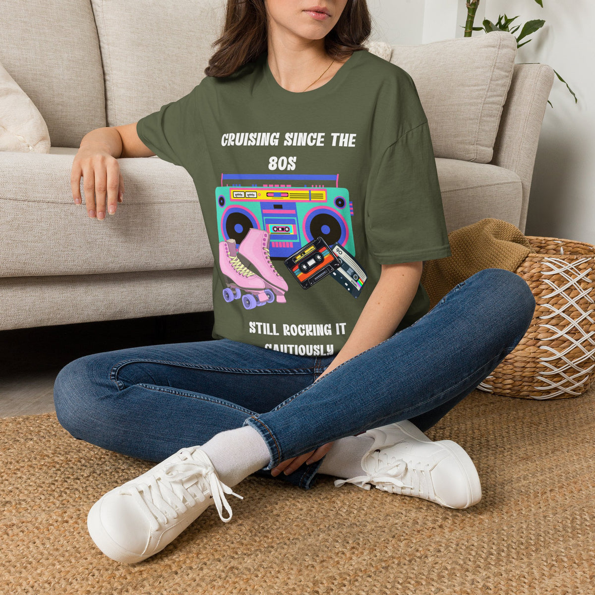 A woman sitting on a woven rug in front of a beige sofa, wearing an olive green graphic t-shirt with a retro 80s boombox and roller skate design, blue jeans, and white sneakers.