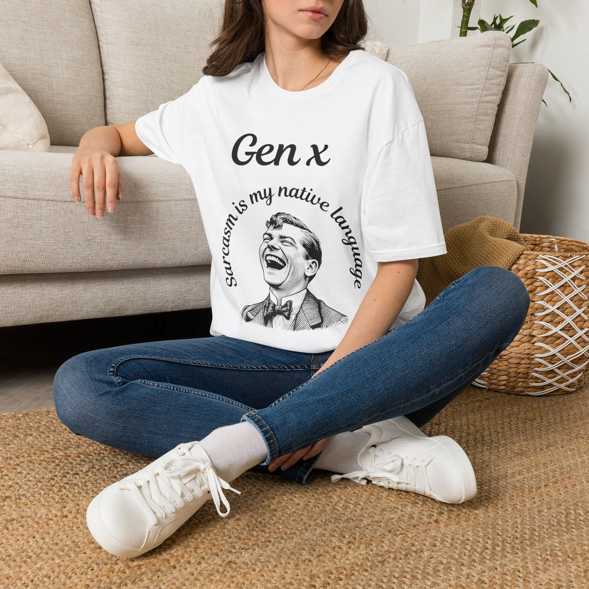 A woman sitting cross-legged on a woven rug in front of a beige sofa, wearing a white graphic T-shirt with black text reading 'Gen x' and 'Sarcasm is my native language' around a laughing man illustration, paired with blue jeans and white sneakers.