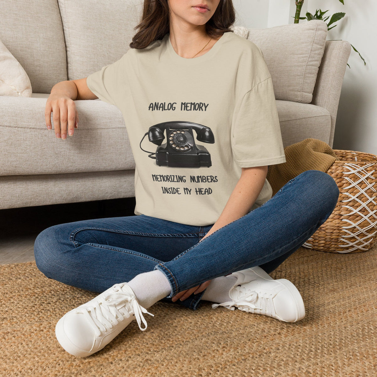A sand short-sleeve graphic T-shirt laid flat on a gray wood-plank background. The shirt has a black printed design featuring a vintage rotary telephone and the text 'ANALOGIC MEMORY' and 'MEMORIZING NUMBERS INSIDE MY HEAD.