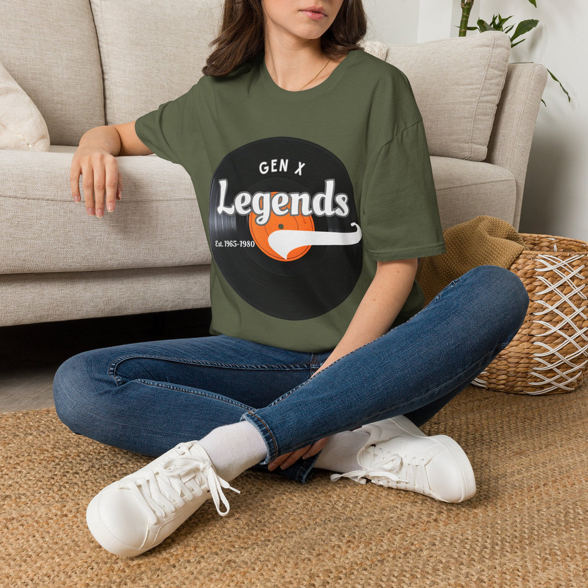 A woman sitting cross-legged on a woven rug in front of a beige sofa, wearing a green graphic T-shirt with a vinyl record design that reads 'GEN X Legends est 1965-1980' 
