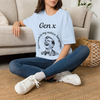 A woman sitting cross-legged on a woven rug in front of a beige sofa, wearing a light blue graphic T-shirt with the text 'Gen x' and 'sarcasm is my native language' around a laughing man illustration, paired with blue jeans and white sneakers.