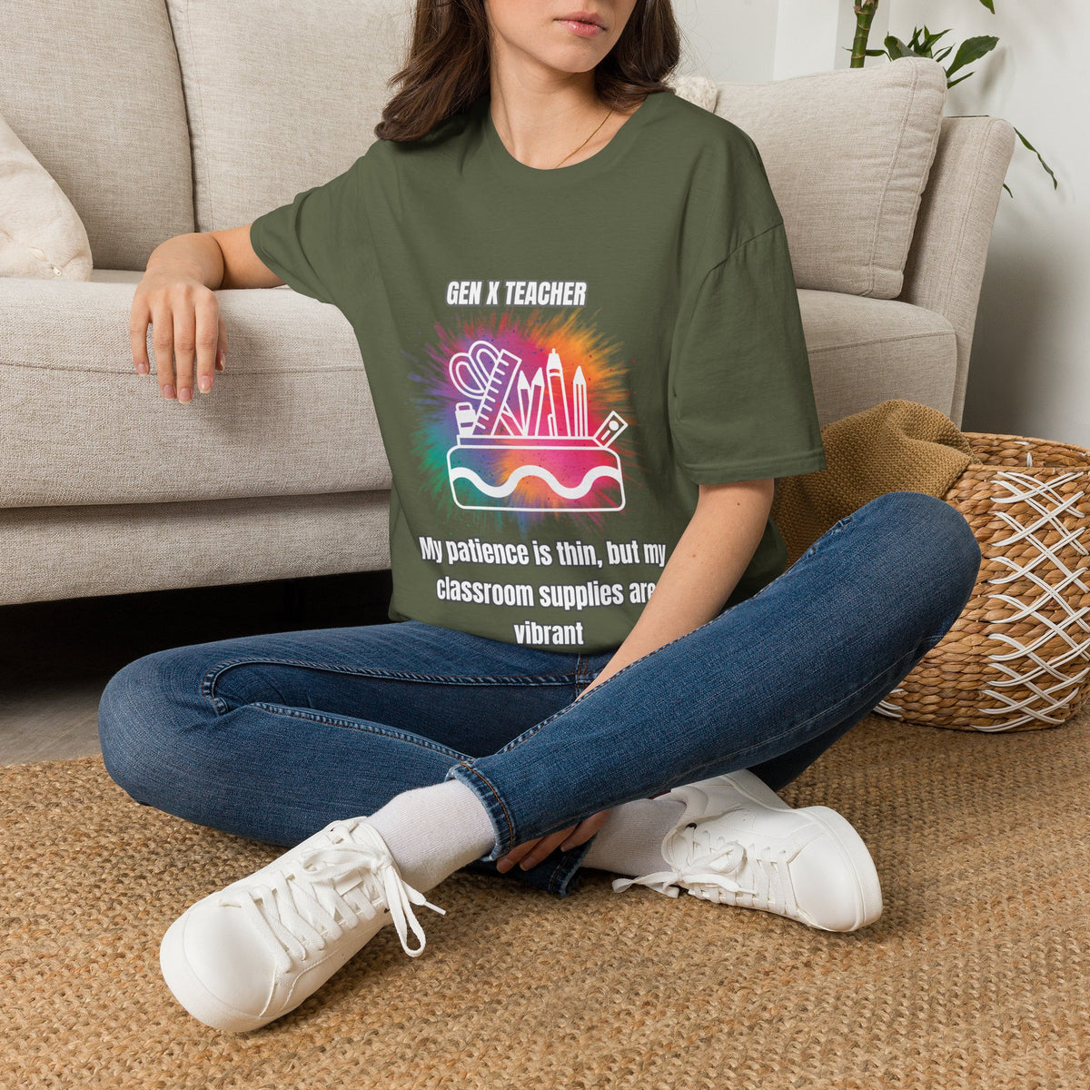 A woman sitting cross-legged on a woven rug in front of a beige sofa, wearing an green graphic T-shirt with a colorful school supplies design and the text 'GEN X TEACHER' and 'My patience is thin, but my classroom supplies are vibrant'.