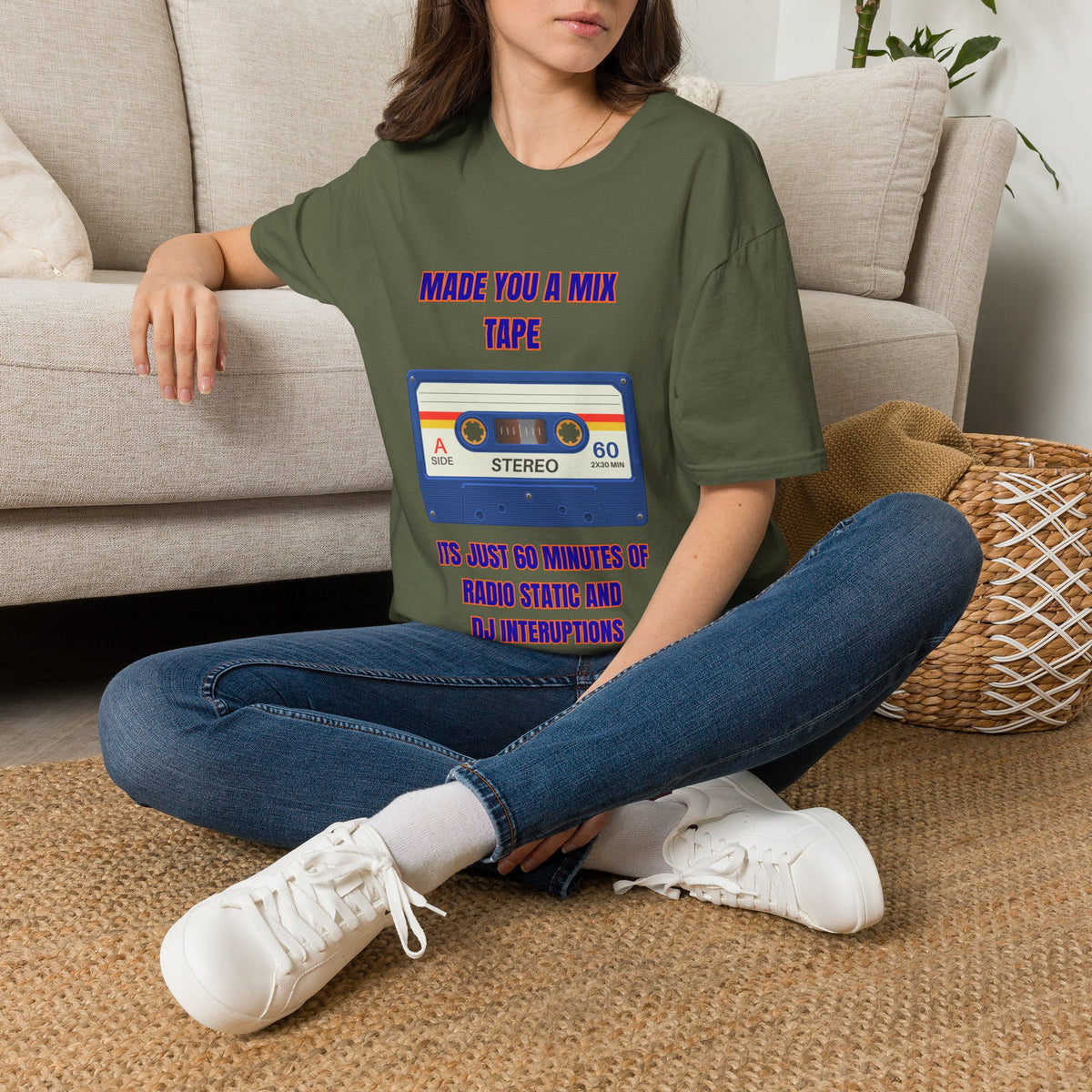 A woman sitting on a woven rug in front of a beige sofa, wearing a green graphic t-shirt with a retro cassette tape design and blue jeans.