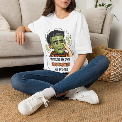 A woman sits cross-legged on a woven rug in front of a beige sofa, wearing an oversized white graphic T-shirt with a child in a halloween mask illustration and the text 'INHALING MY OWN CONFUSION ALL EVENING' paired with blue jeans and white sneakers.