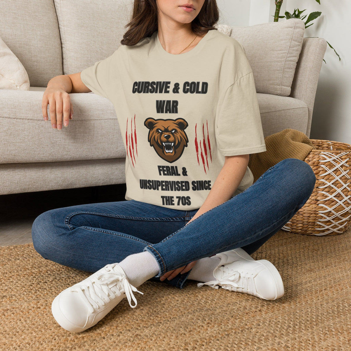 A woman sitting cross-legged on a woven rug in front of a light beige sofa, wearing a sand graphic t-shirt with a bear design and text, blue jeans, and white sneakers.