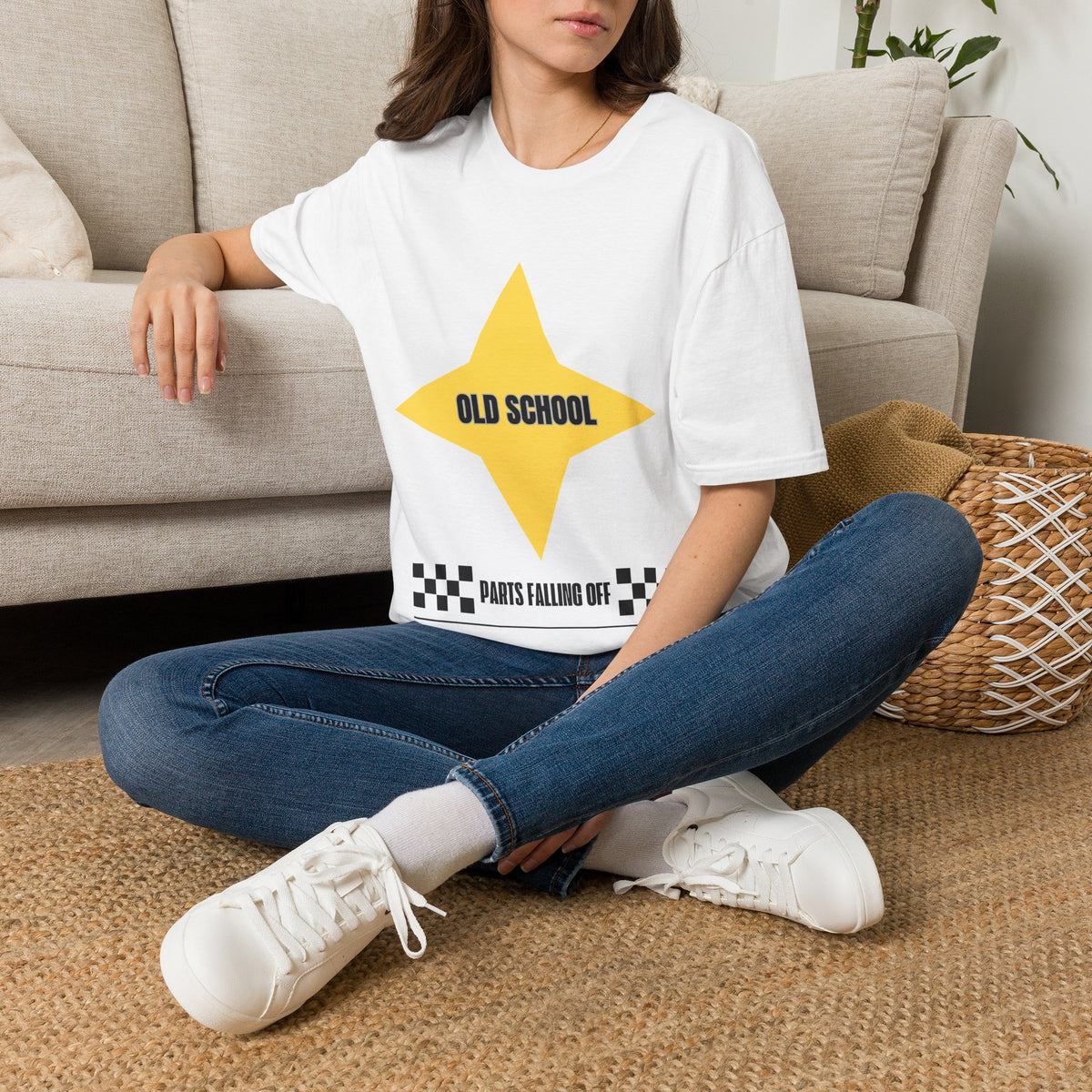 A woman sitting cross-legged on a woven rug in front of a beige sofa, wearing an oversized white graphic T-shirt with a yellow star and the text 'OLD SCHOOL' and 'PARTS FALLING OFF', paired with blue jeans and white sneakers.