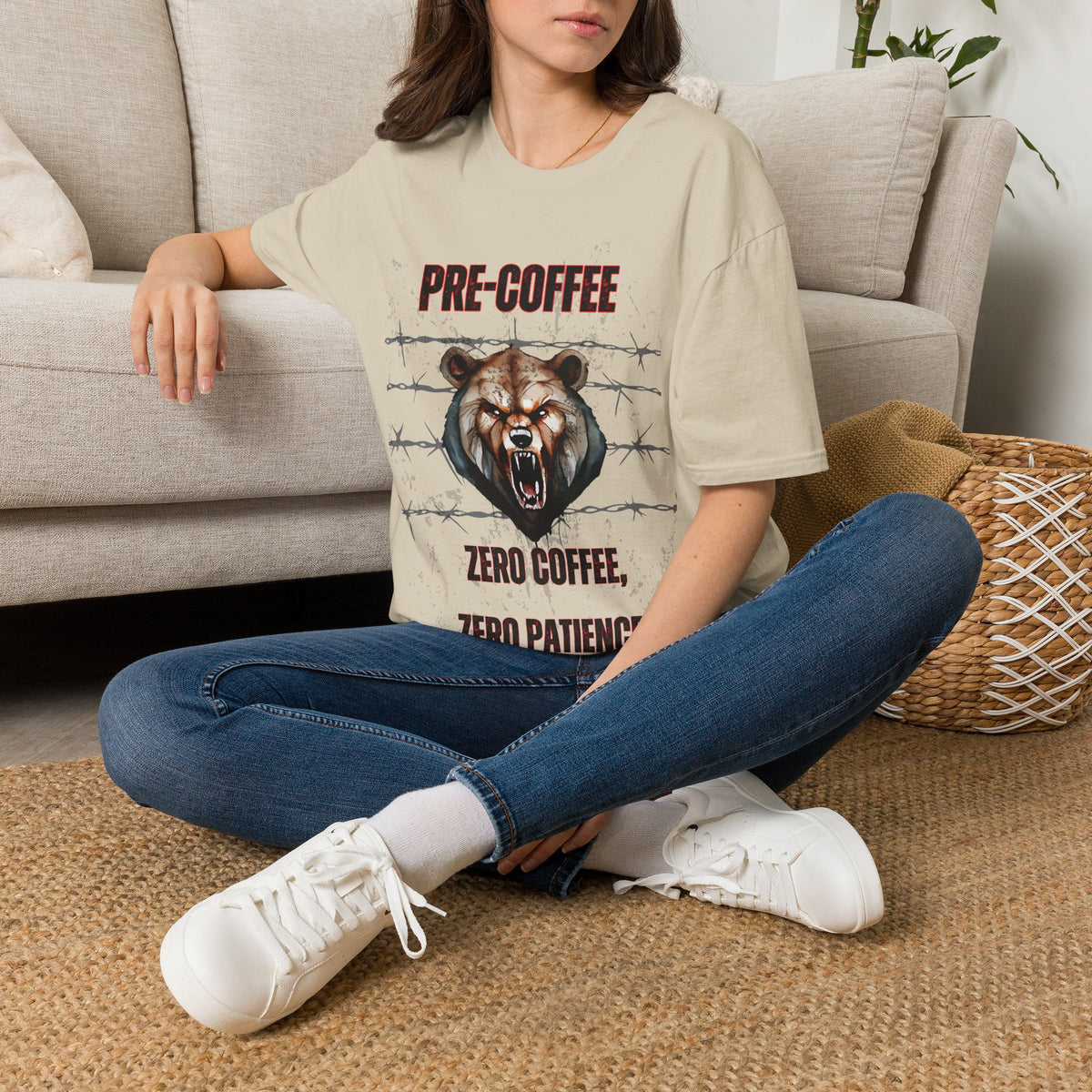 A woman sitting on a woven rug in front of a beige sofa, wearing a beige graphic t-shirt with a bear design and the text 'PRE-COFFEE ZERO COFFEE ZERO PATIENCE', blue jeans, and white sneakers.