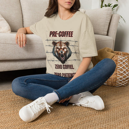 A woman sitting on a woven rug in front of a beige sofa, wearing a beige graphic t-shirt with a bear design and the text 'PRE-COFFEE ZERO COFFEE ZERO PATIENCE', blue jeans, and white sneakers.