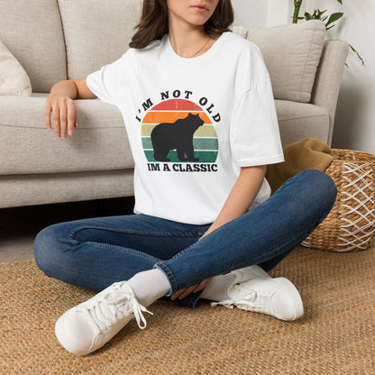 A woman sitting cross-legged on a woven rug in front of a beige sofa, wearing a white graphic T-shirt with a bear silhouette and the text 'I'M NOT OLD I'M A CLASSIC' over a retro sunset design, paired with blue jeans and white sneakers.