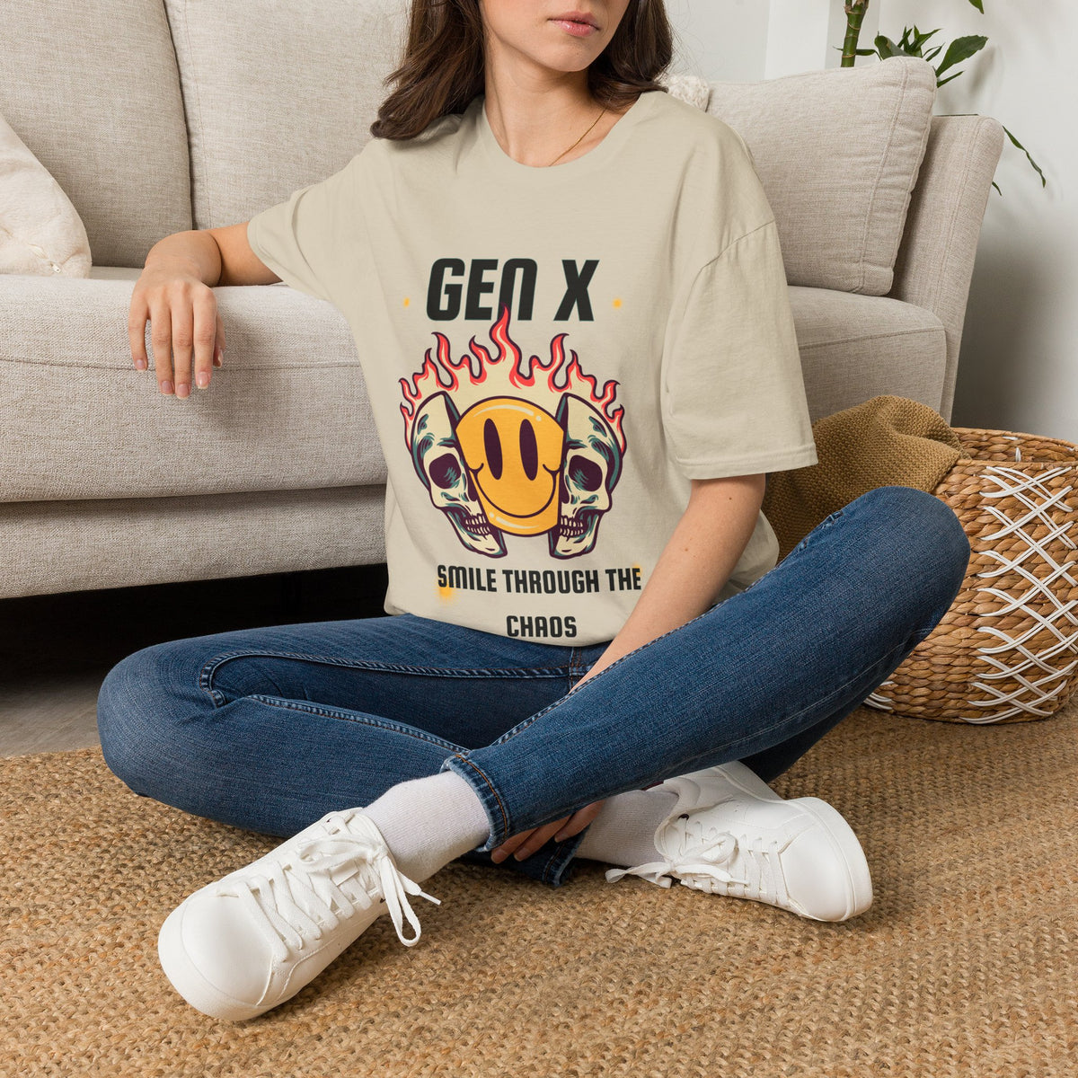 A woman sat on a rug next to the couch wearing a sand graphic t-shirt, featuring a design with two skulls in flames and a yellow smiley face. The text reads 'GEN X SMILE THROUGH THE CHAOS'.