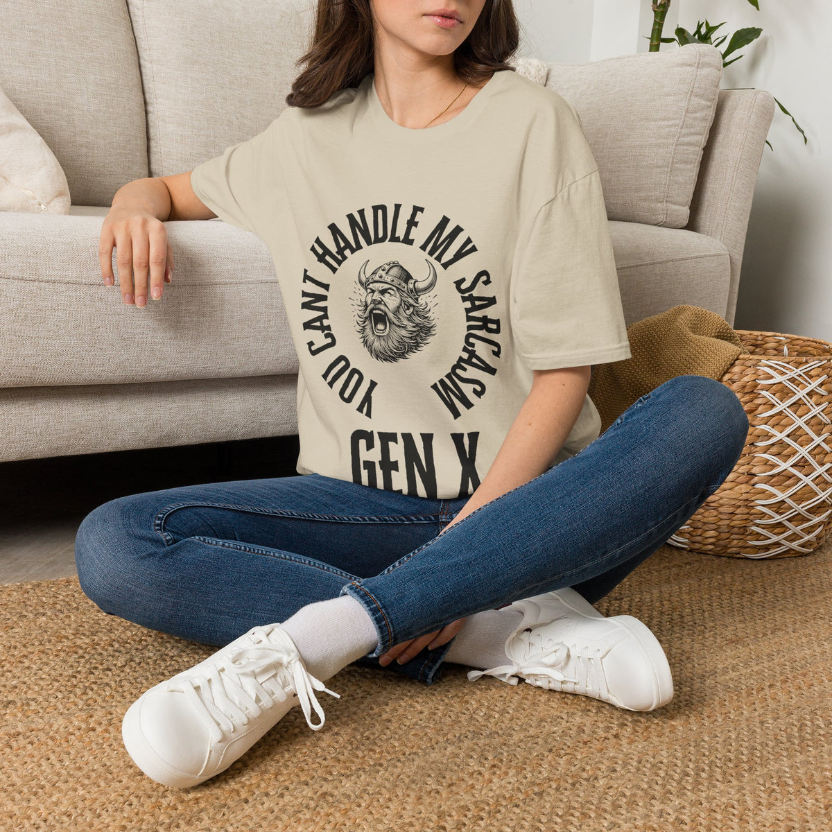 A woman sat on a rug next to a couch wearing a white short-sleeve graphic t-shirt with a black circular print featuring a roaring viking man and the text 'YOU CAN'T HANDLE MY SARCASM GEN X'. 