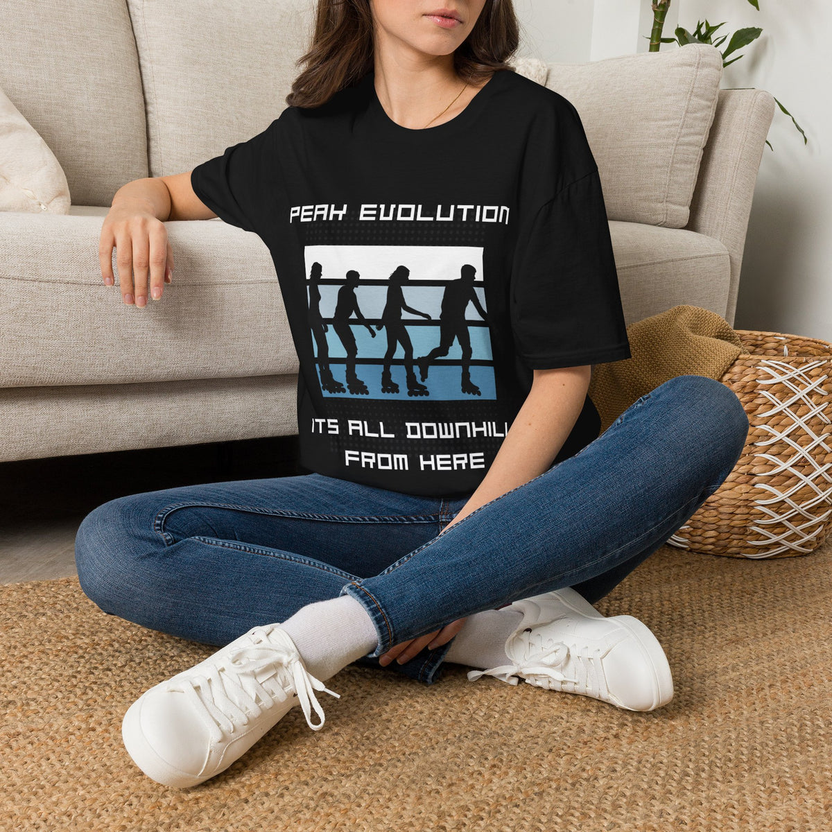 A woman sitting cross-legged on a woven rug in front of a beige sofa, wearing a black graphic T-shirt with the text 'PEAK EVOLUTION IT'S ALL DOWNHILL FROM HERE' and a white, blue, and black boxing-ring graphic.