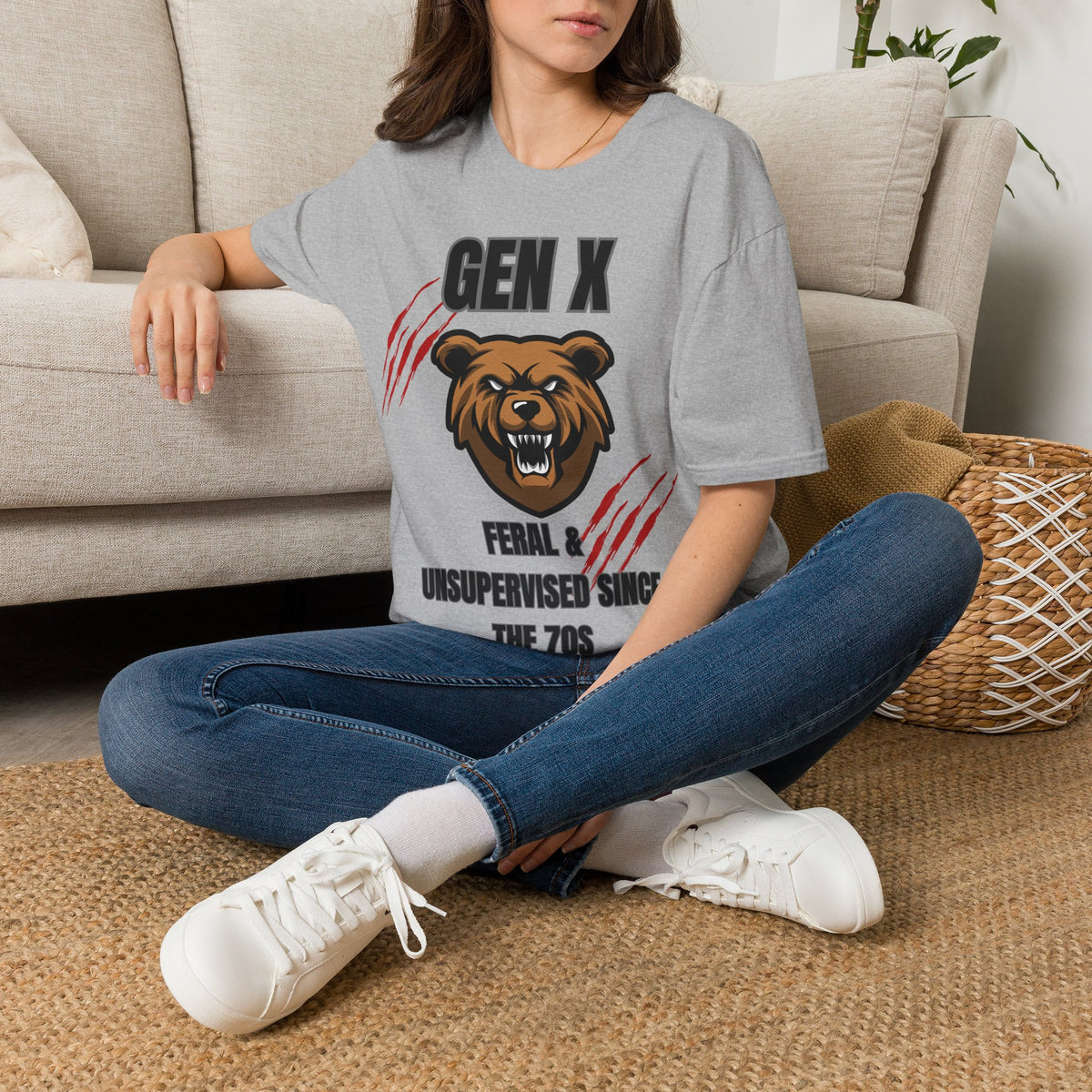 A woman sitting cross-legged on a woven rug in front of a beige sofa, wearing a gray graphic T-shirt with a bear head and the text 'GEN X FERAL & UNSUPERVISED SING THE 7OS'.