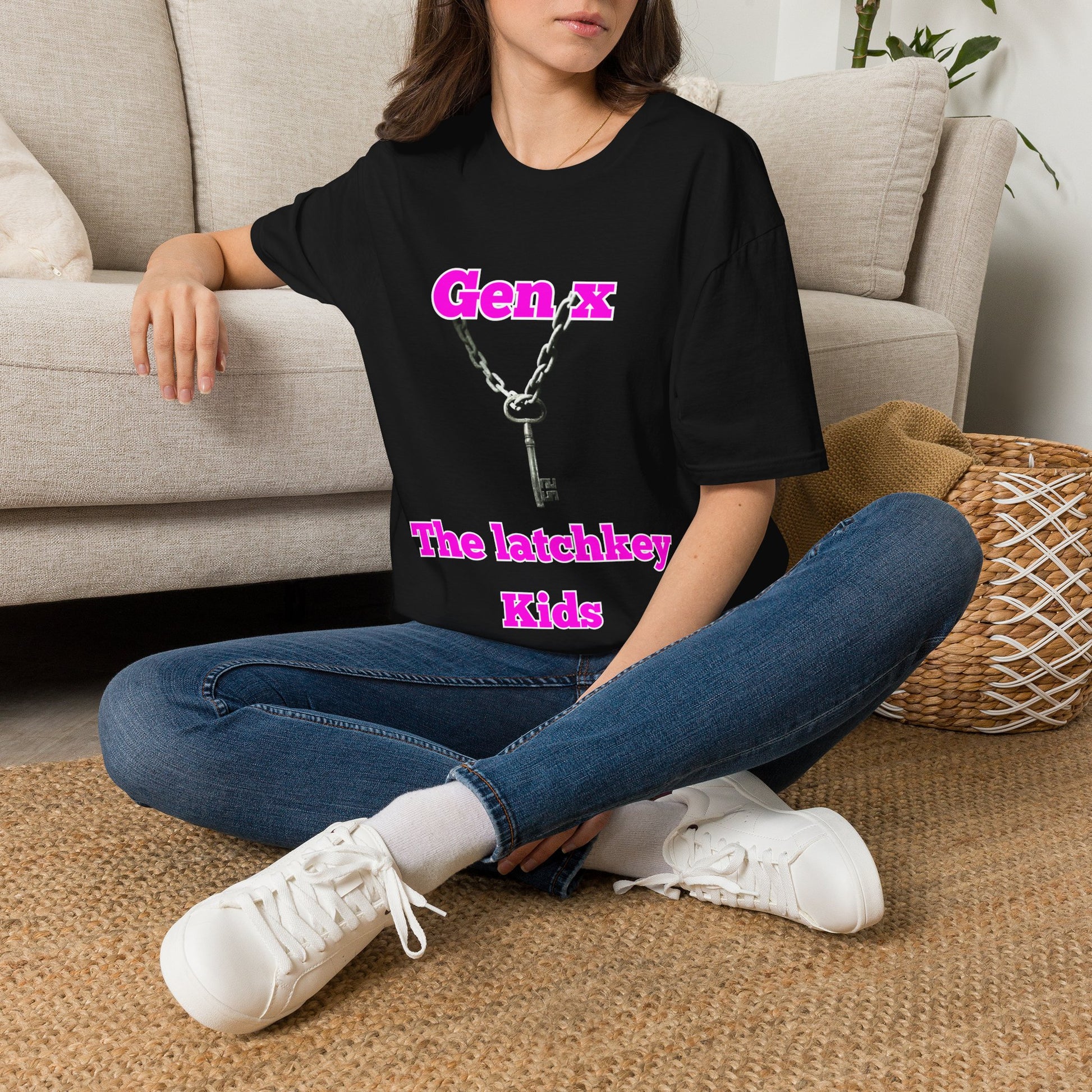 A woman sitting cross-legged on a woven rug in front of a beige sofa, wearing an oversized black graphic T-shirt with pink text reading 'Gen X The latchkey Kids' and a silver key pendant graphic.