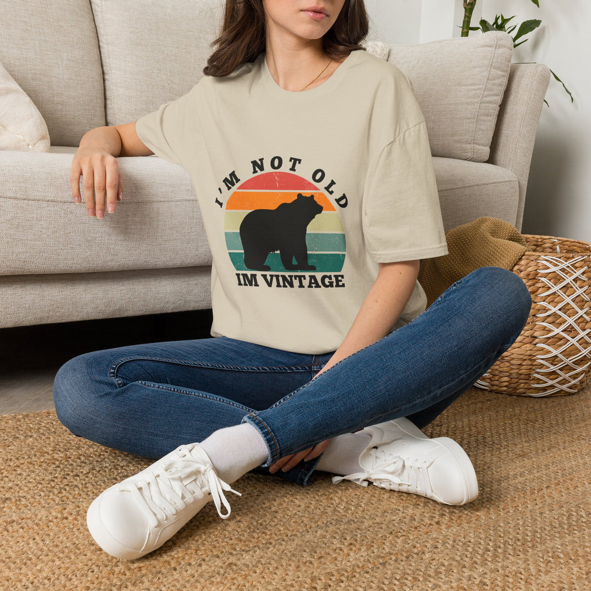 A woman sitting cross-legged on a woven rug in front of a beige sofa, wearing an oversized sand graphic T-shirt with a retro bear design and the text 'I'M NOT OLD I'M VINTAGE'.
