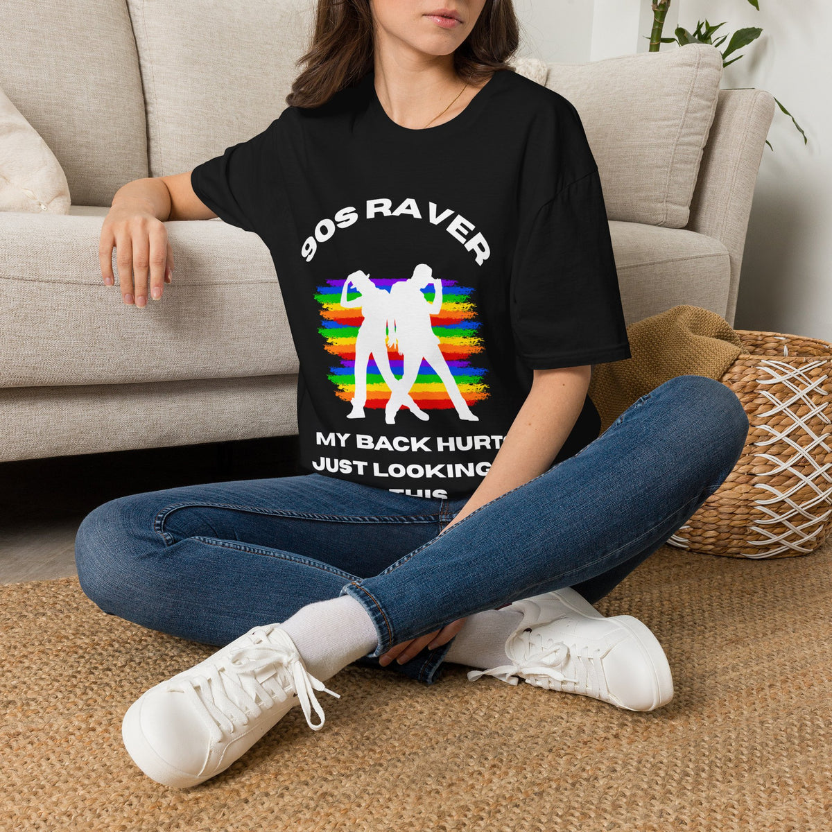 A woman sitting cross-legged on a woven rug in front of a beige sofa, wearing a black graphic T-shirt with white text and a rainbow-striped retro figure design, blue jeans, and white sneakers.