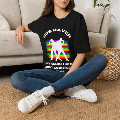 A woman sitting cross-legged on a woven rug in front of a beige sofa, wearing a black graphic T-shirt with white text and a rainbow-striped retro figure design, blue jeans, and white sneakers.