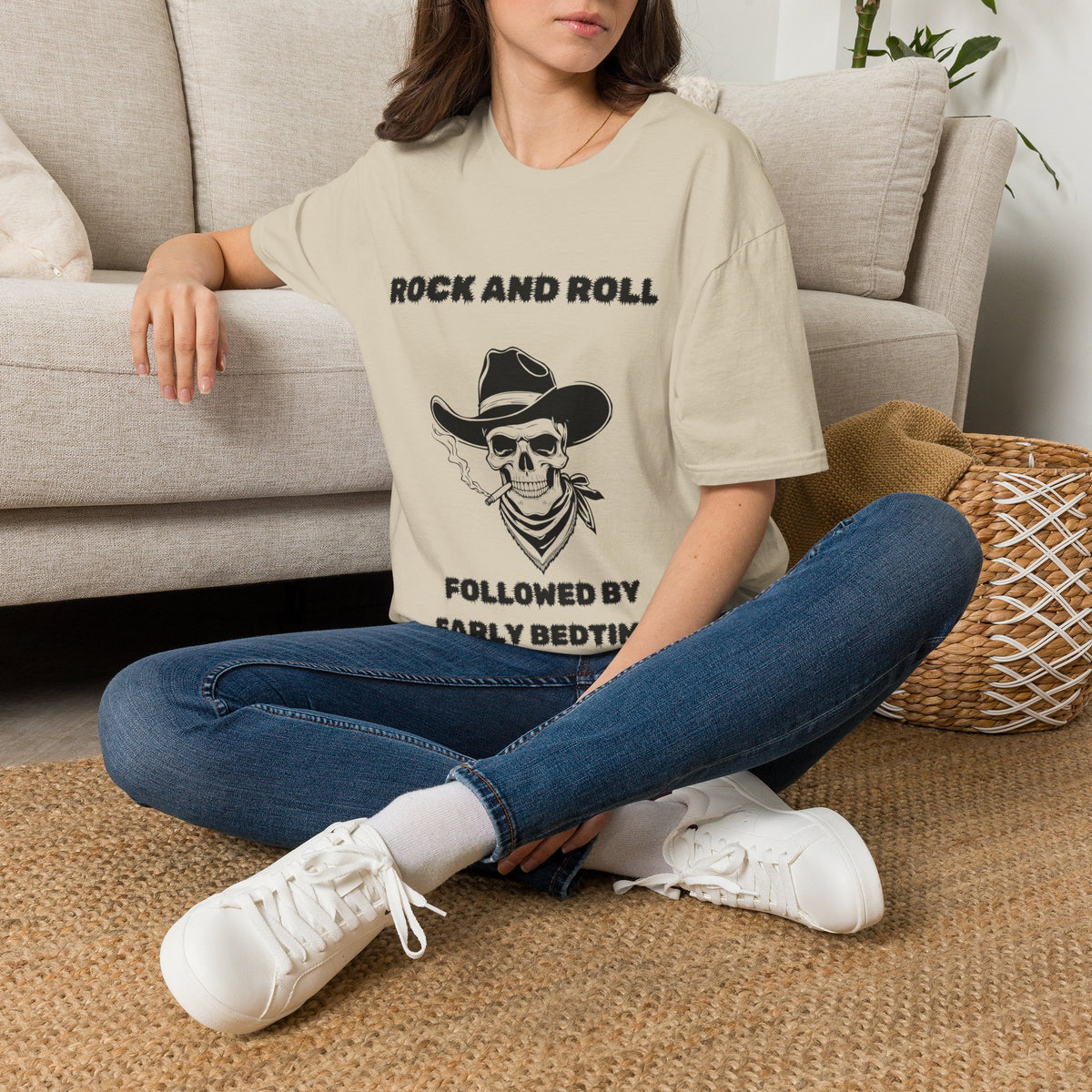 A woman sits cross-legged on a woven rug in front of a light beige sofa, wearing an oversized sand graphic T-shirt with a black skull-and-cowboy print and text, blue jeans, and white sneakers.