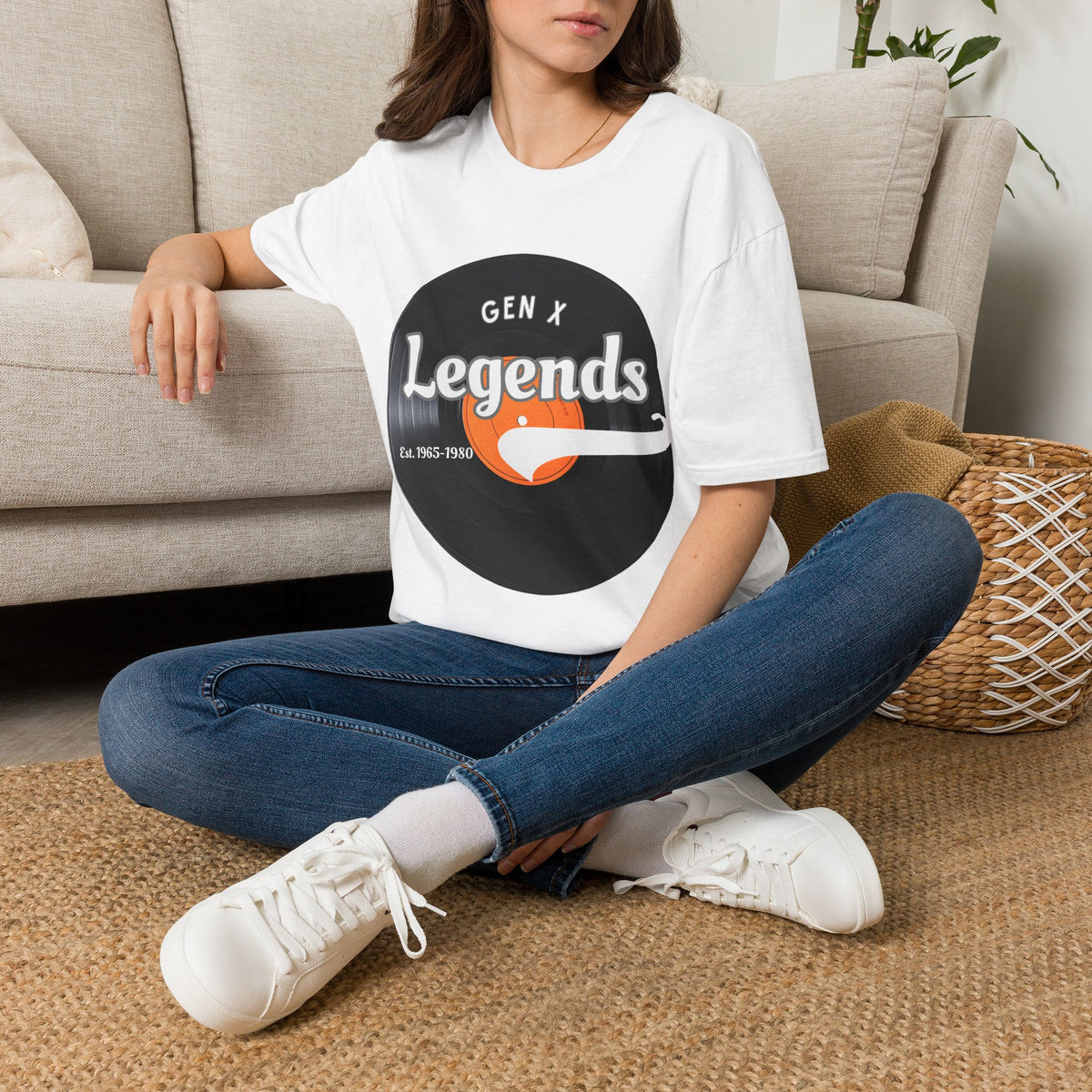 A woman sitting cross-legged on a woven rug in front of a beige sofa, wearing a white graphic T-shirt with a vinyl record that reads 'GEN X Legends est 1965-1980' 