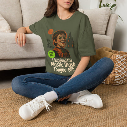 A woman sitting cross-legged on a woven rug in front of a beige sofa, wearing a green graphic T-shirt with a large vintage-style illustration of a child in halloween mask and the text 'I SURVIVED THE PLASTIC MASK SLIT'.
