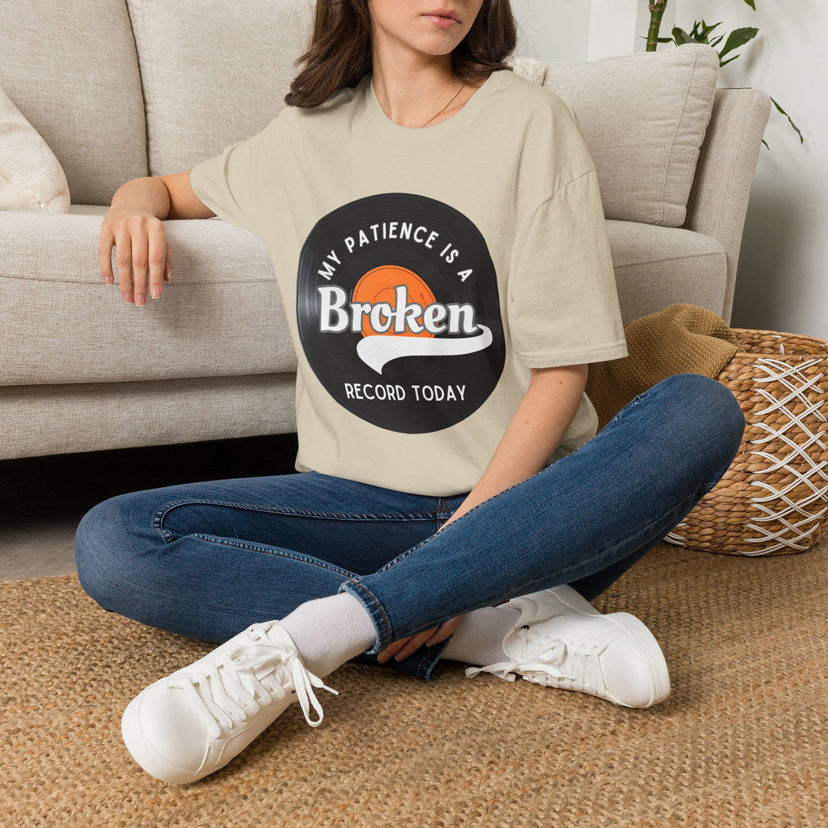 A woman sitting cross-legged on a woven rug in front of a beige sofa, wearing an oversized sand T-shirt with a vinyl record graphic that reads 'MY PATIENCE IS A BROKEN RECORD TODAY' and blue jeans with white sneakers