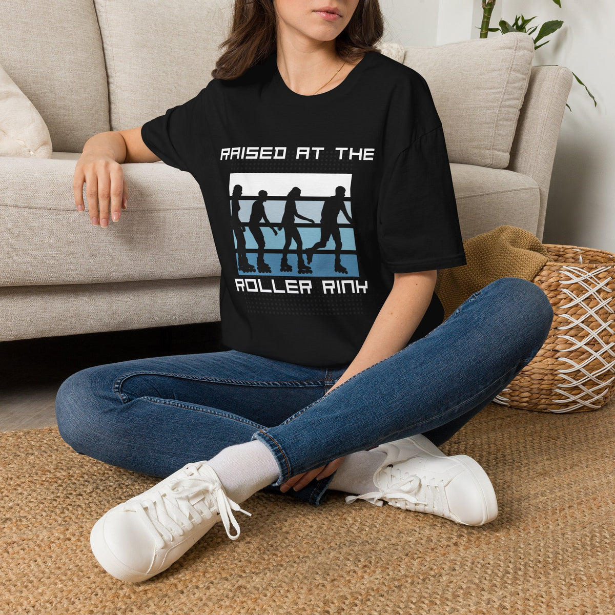 A woman sitting cross-legged on a woven rug in front of a beige sofa, wearing a black graphic T-shirt with the text 'RAISED AT THE ROLLER RINK' and a white, blue, and black roller skate design.