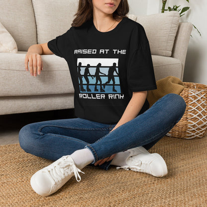 A woman sitting cross-legged on a woven rug in front of a beige sofa, wearing a black graphic T-shirt with the text 'RAISED AT THE ROLLER RINK' and a white, blue, and black roller skate design.