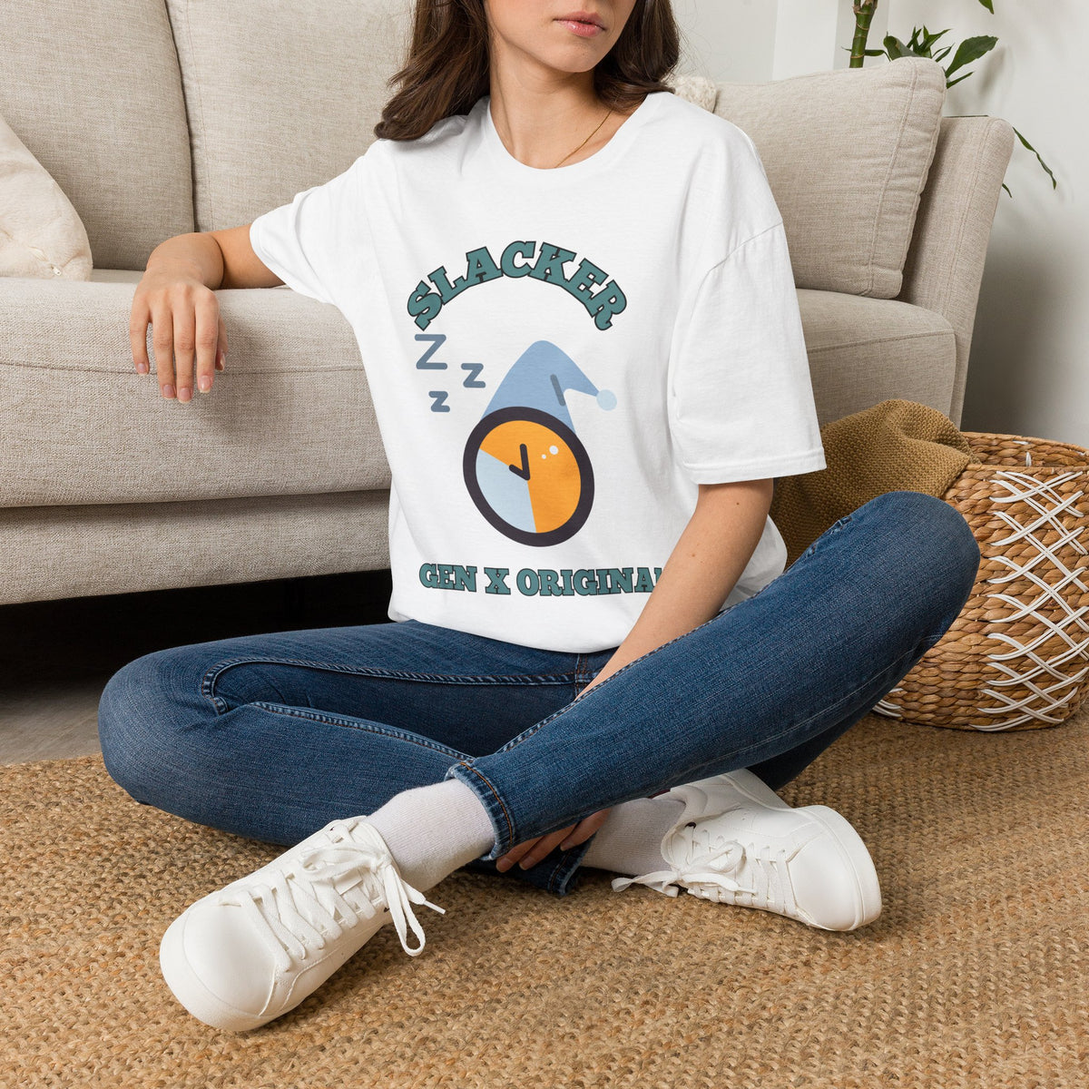 A woman sitting cross-legged on a woven rug in front of a beige sofa, wearing a white graphic T-shirt with the text 'he can't suck himself' and a red arrow graphic, paired with blue jeans and white sneakers.