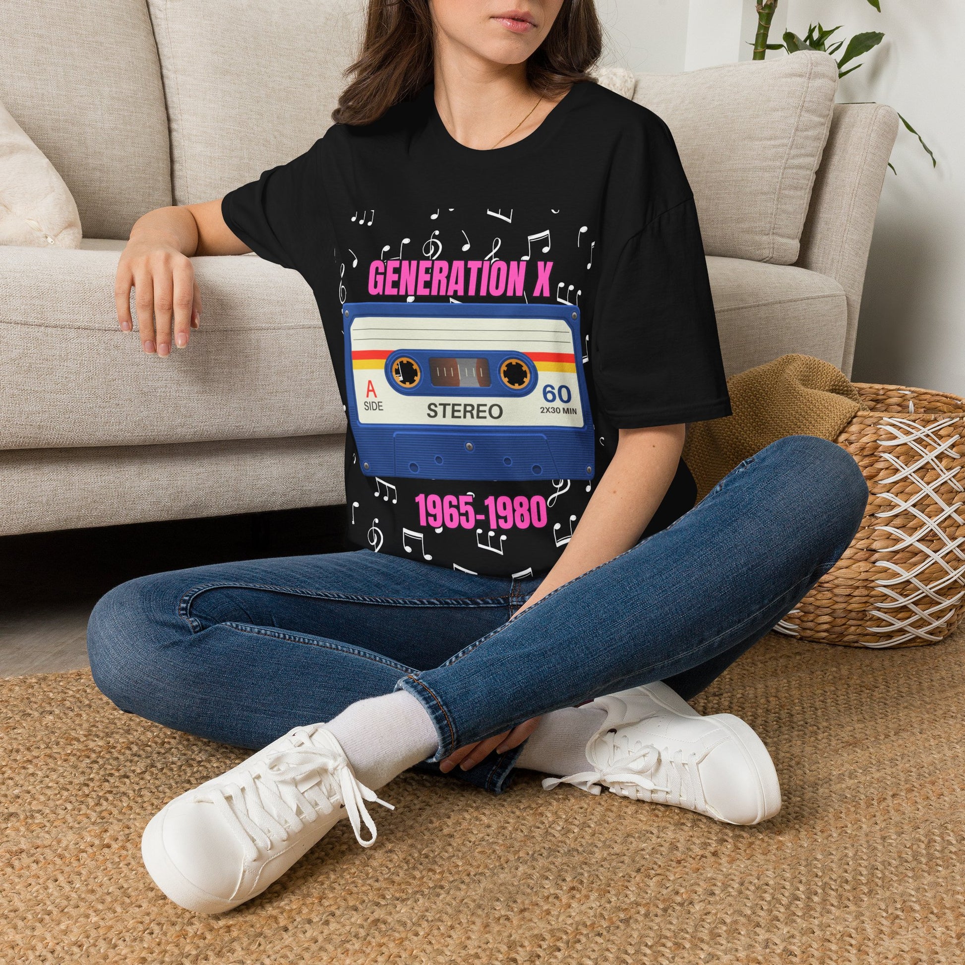 A woman sitting cross-legged on a woven rug in front of a beige sofa, wearing a black graphic T-shirt with a retro cassette tape design and the pink text 'GENERATION X 1965-1980'.