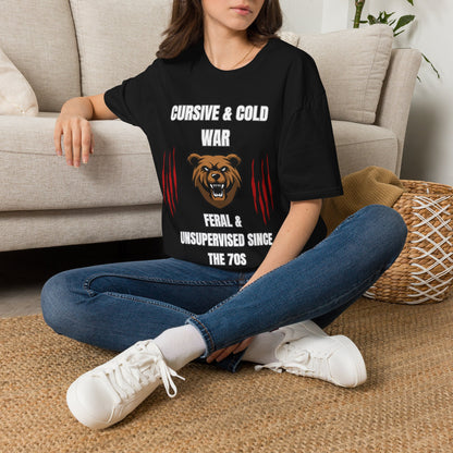 A woman sitting on a woven rug in front of a beige couch, wearing a black graphic t-shirt with a bear design and the text 'CURSIVE & COLD WAR FERAL & UNSUPERVISED SINCE THE 70S'.