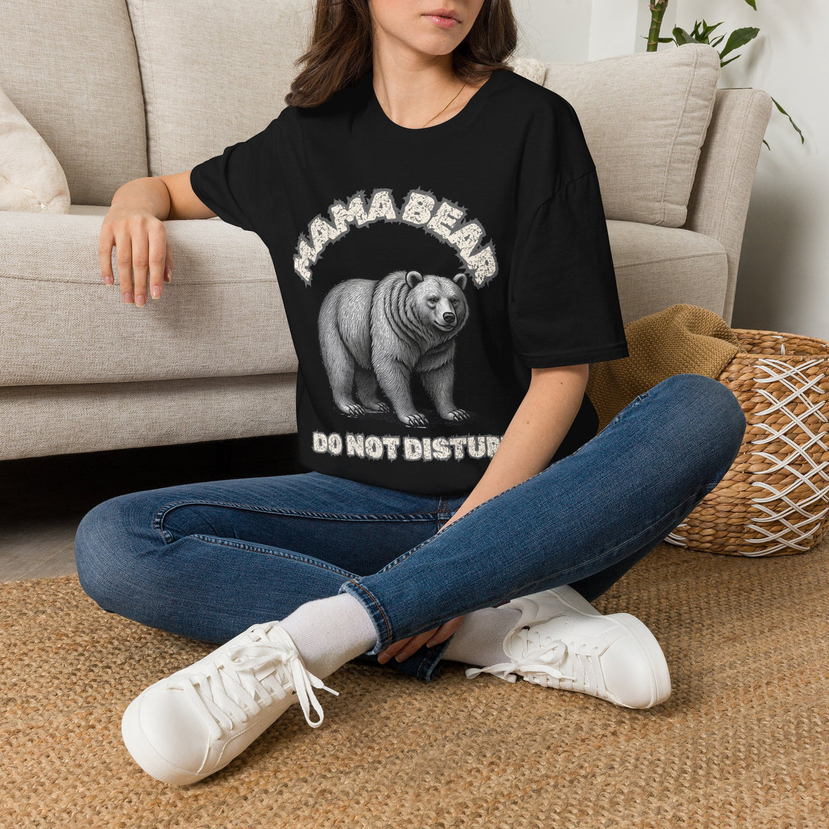 A woman sitting on a woven rug in front of a beige sofa, wearing a black graphic t-shirt with a white bear print and the text 'MAMA BEAR DO NOT DISTURB', paired with blue jeans and white sneakers.
