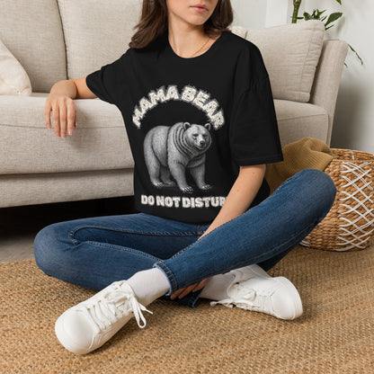 A woman sitting on a woven rug in front of a beige sofa, wearing a black graphic t-shirt with a white bear print and the text 'MAMA BEAR DO NOT DISTURB', paired with blue jeans and white sneakers.